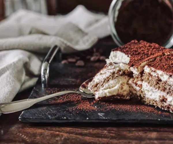 Table de trois restaurants Bordeaux, part de tiramisu saupoudrée de cacao posée sur une ardoise noire avec une cuillère, décor de grains de café en arrière plan